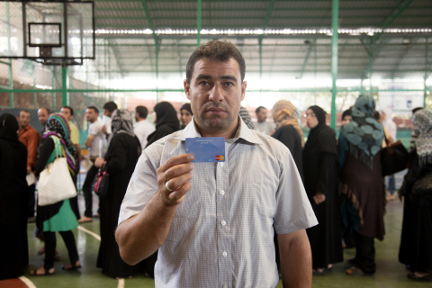 敘利亞難民Ali Ahmad Farhat拿著他的萬事達卡/WFP電子卡，其他人也在籃球場排隊領取自己的卡。（照片：美國商業資訊）